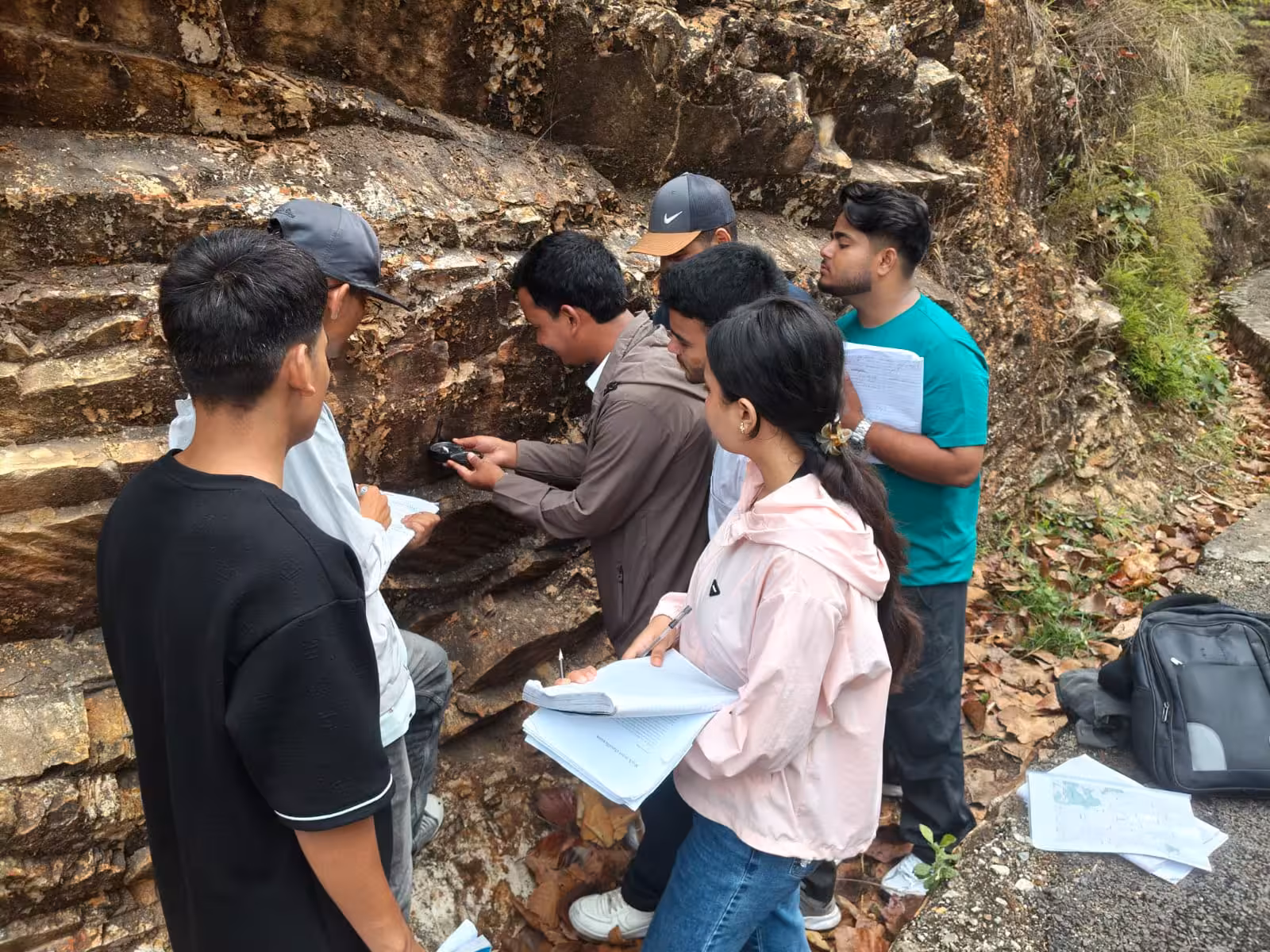 Students taking notes at an exposed rock face, one holding a clipboard, packs on the ground