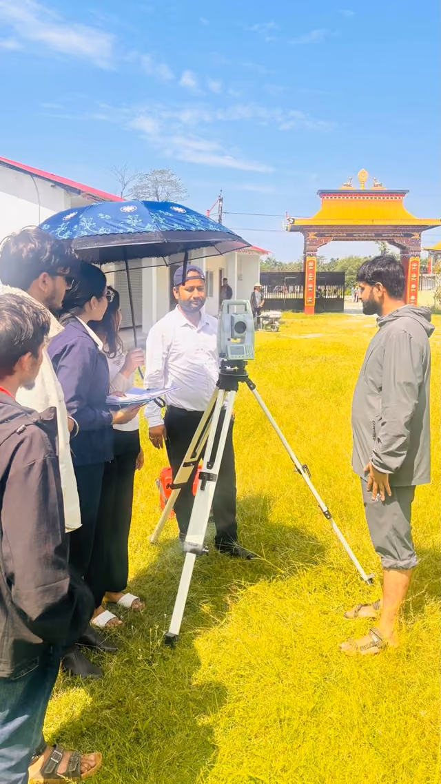 A group round a survey tripod on the lawn, the campus gateway behind them