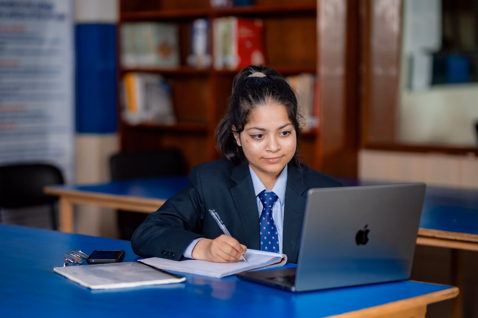 A student writing beside her laptop in the reading room, bookshelves and framed certificates behind