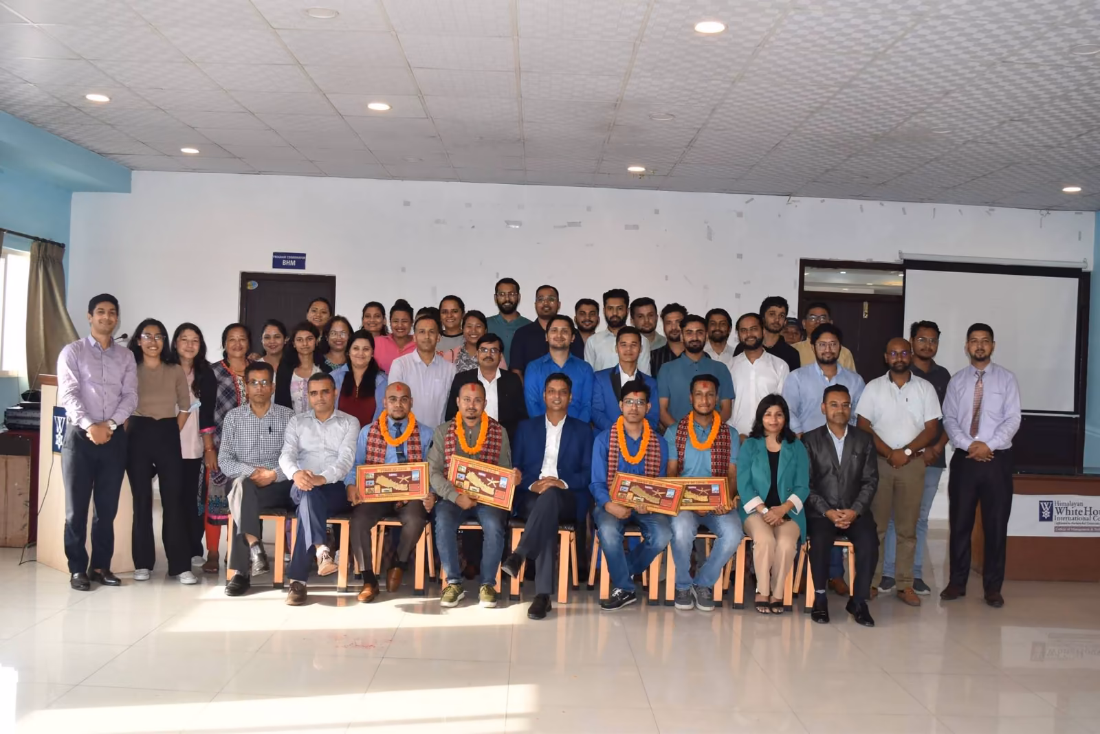 Staff and students in rows across a hall, several of them holding framed certificates