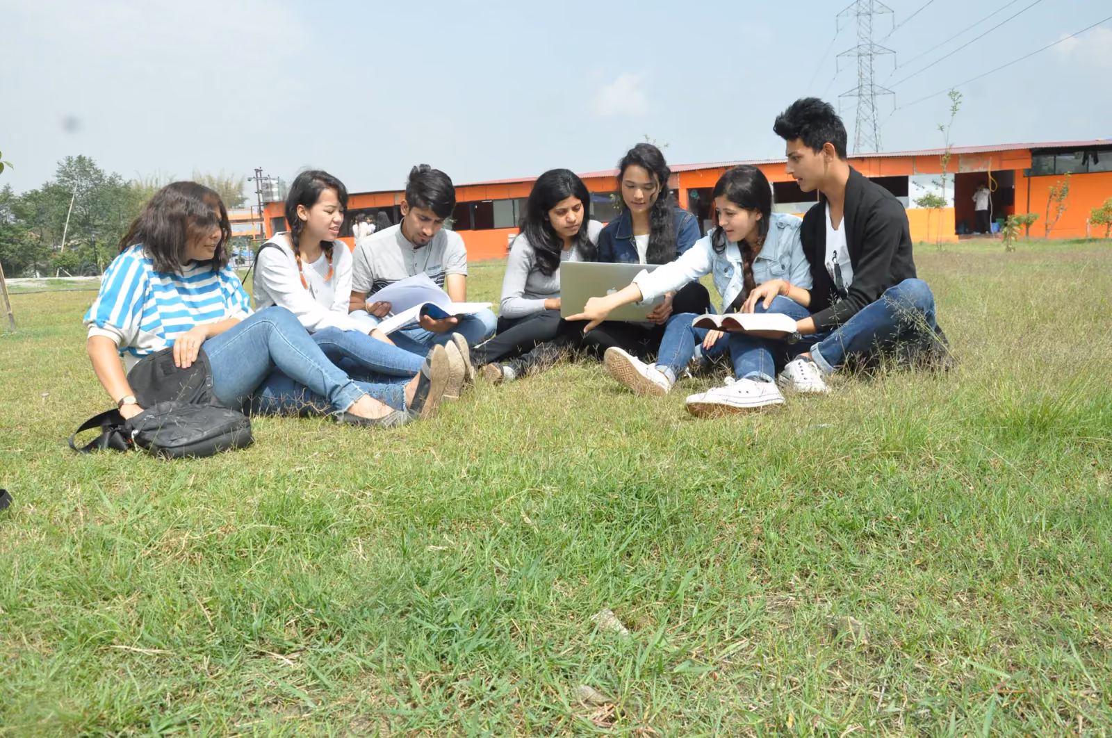 Nine students sitting on the grass with books and a laptop, the campus block behind