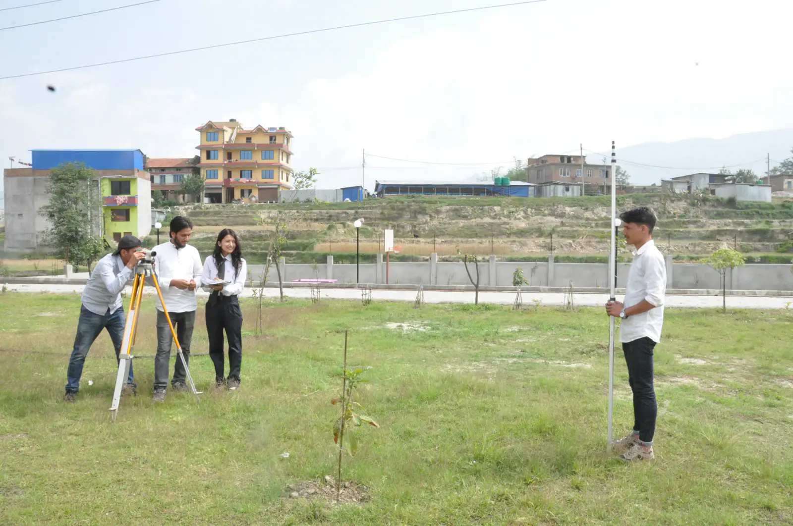 Students setting up a survey tripod on open ground, the college buildings across the field