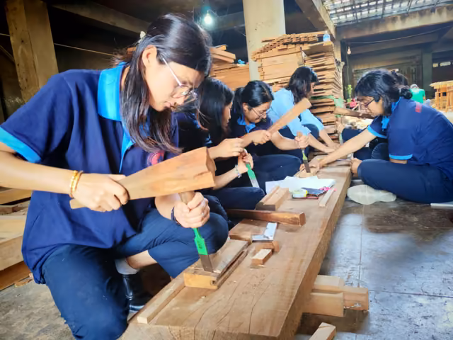 Students in blue overalls planing and chiselling timber at a low bench in the wood workshop