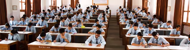 Students at single desks through a long examination hall, curtains drawn along the windows