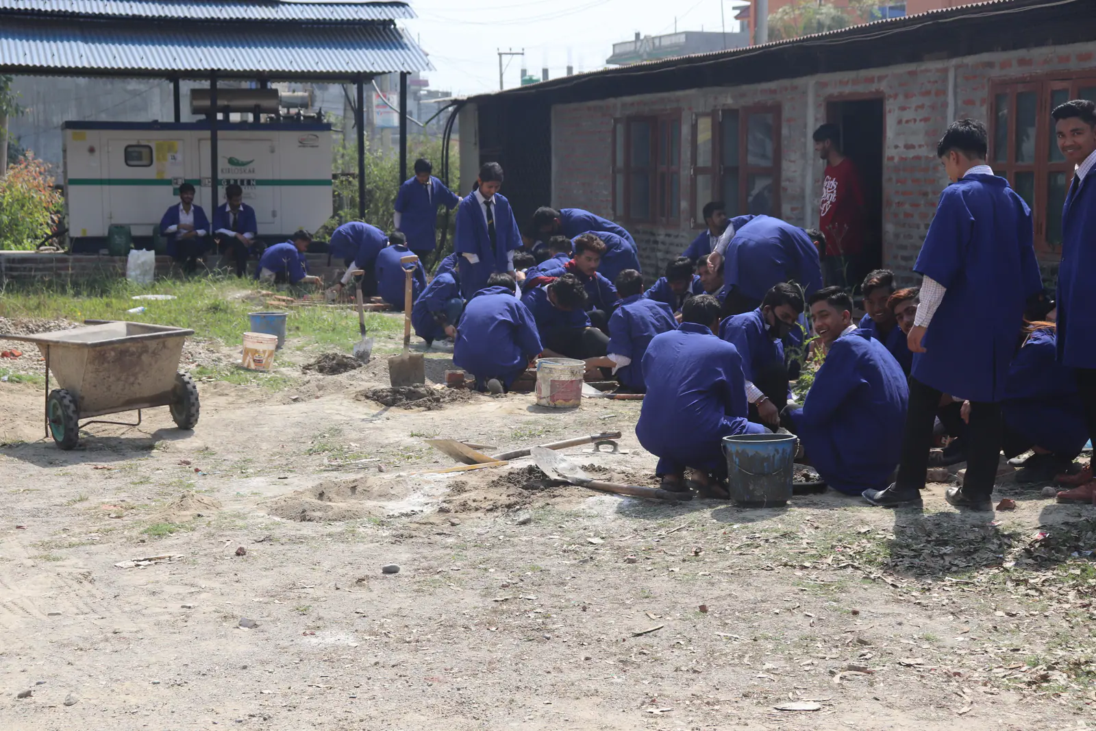Students in blue overalls at work on the ground outside the workshop shed, a concrete mixer beside them