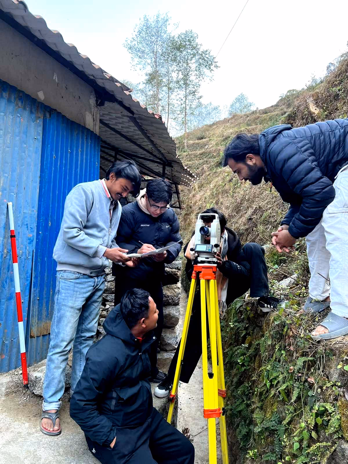 Students crouched over a levelling instrument beside a blue shed, the staff held on the slope