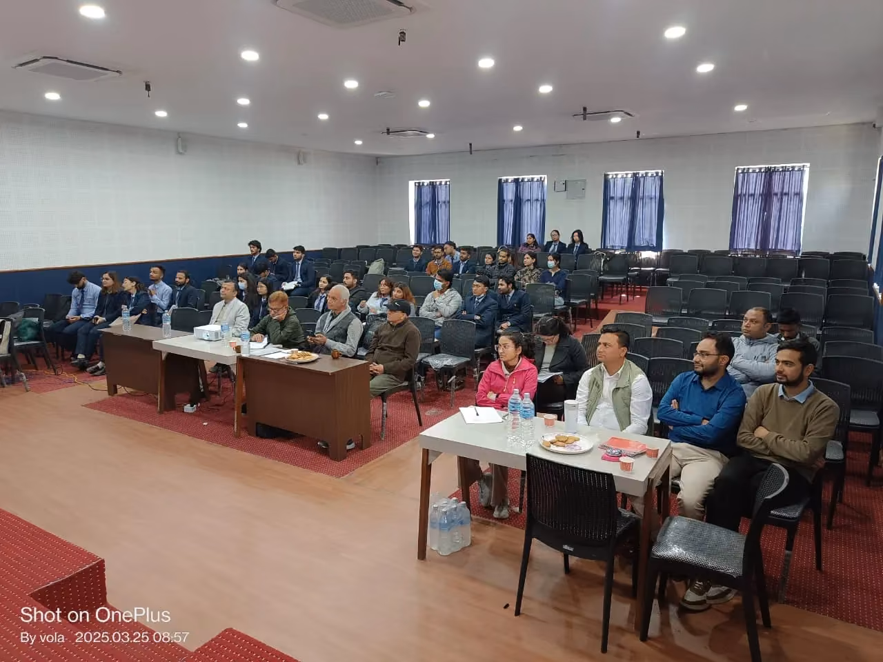 An audience seated in a seminar room, a panel at the table in front