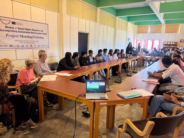 Participants round a long conference table under a project meeting banner, laptops open