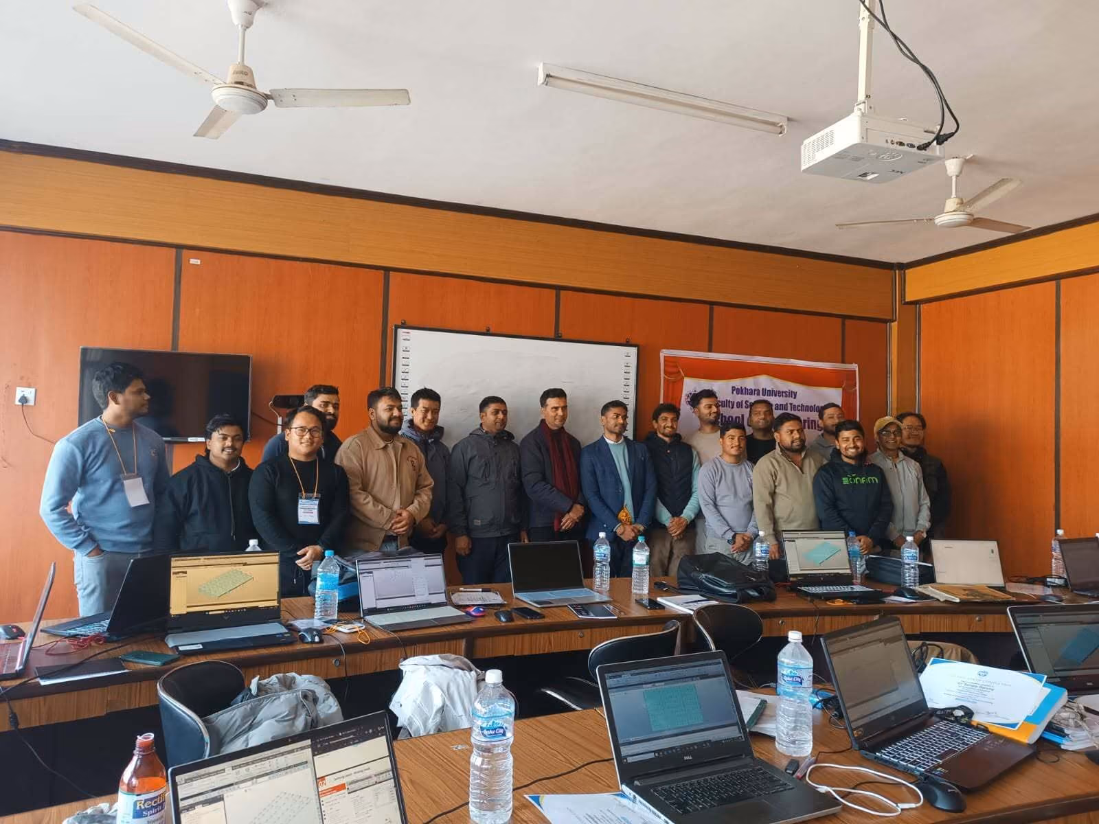 Participants at laptops round the long tables of a training room, a projector screen at the end