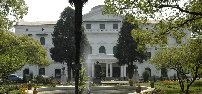 The campus's white colonnaded building behind its lawn trees, cars parked along the front