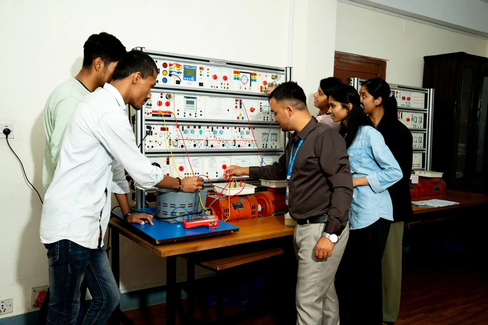 Students at a wall of electrical trainer panels, one connecting leads at the bench