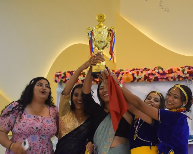 Five students lifting a trophy together in front of a decorated backdrop
